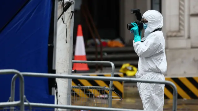 technician wearing white protective clothing, a blue face mask and aqua gloves. She is holding a camera to her face to take a photo.