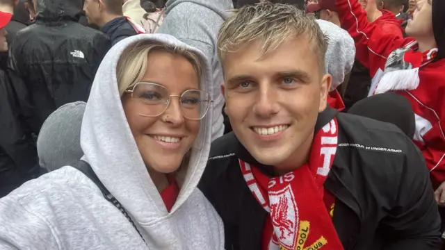 Abbie on the left and Jack on the right posing for a selfie at the parade. Abbie is wearing a grey hoodie with the hood up and glasses and is smiling. Jack is wearing a black jacket and Liverpool FC scarf around his neck. He is smiling too