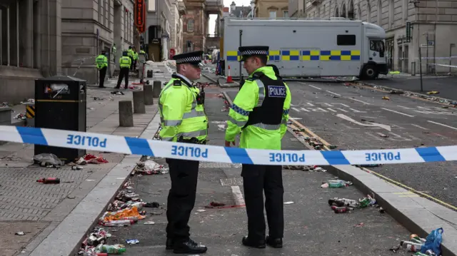 Police stand behind police tape, with debris strewn across the floor