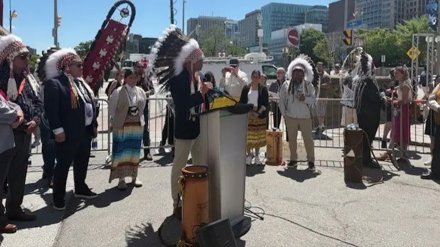 A man in a traditional headress speaks at a lectern, while other stand around him