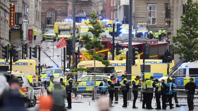 A view of the street after the incident happened, with a large number of police vehicles and ambulances, with lots of people in high vis guarding a fence in front of the street