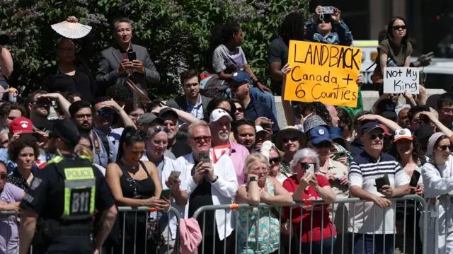 A sign which says 'Landback. Canada + six counties' and another sign which says 'Not my King' held up in the crowd