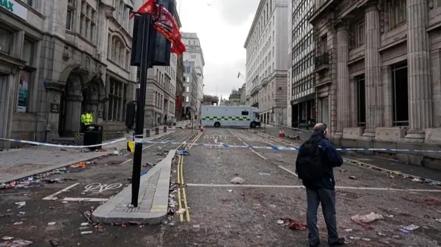 Man stands on a street littered with debris, a police van is in the background
