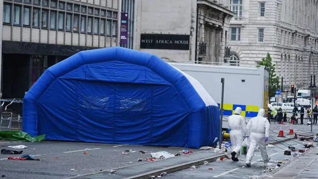 A forensics tent in the street, while two people wearing forensics suits walk past, with the police cordon in the background at the end of the street
