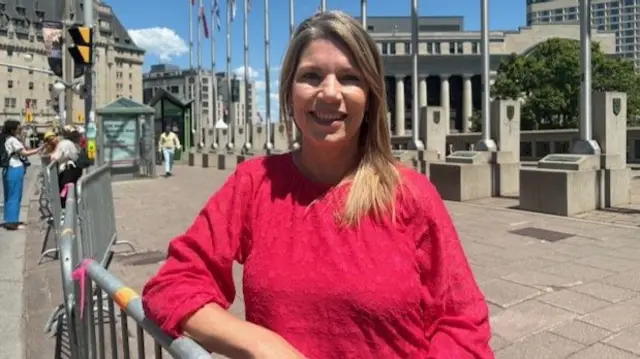 Heather Mitchell, a  blonde woman wearing a red blouse, smiles at the camera from donwtown Ottawa