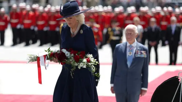 Queen Camila lays flowers, while Charles can be seen standing at a distance behind her
