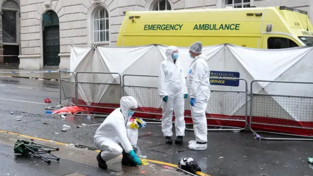 A forensic worker kneels down to collect evidence as two other stand behind. An ambulance, shielded by a white screen, is behind them.