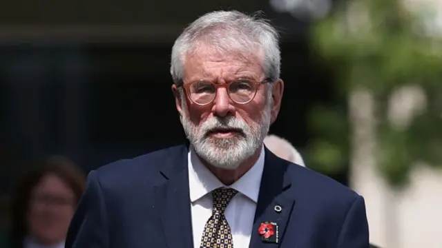 alt="Gerry Adams is on a street. He has white hair and a beard. He's wearing orange glasses and a navy suit with a white shirt and a tie.