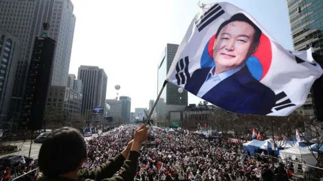 Backview of a woman waving a large South Korean flag with a photo of Yoon Suk Yeol printed over it. She's standing before a large crowd gathered on a big road.