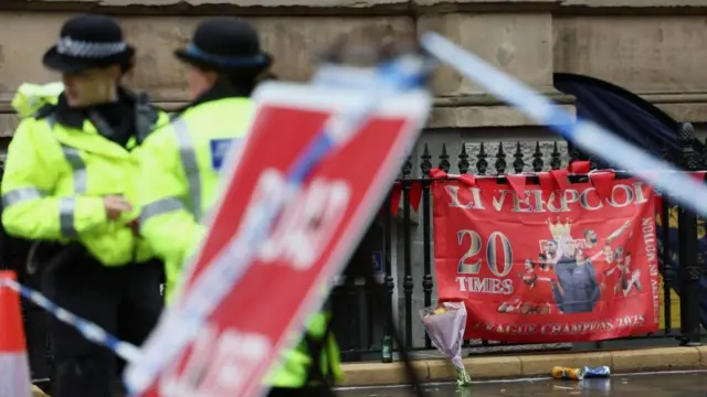 Two police officers next to a road closed sign, near a fence adorned with a Liverpool FC flag