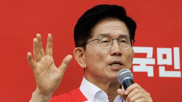 Kim Moon-soo speaking into a microphone while holding his other hand up. He is wearing round glasses and standing in front of a red background.