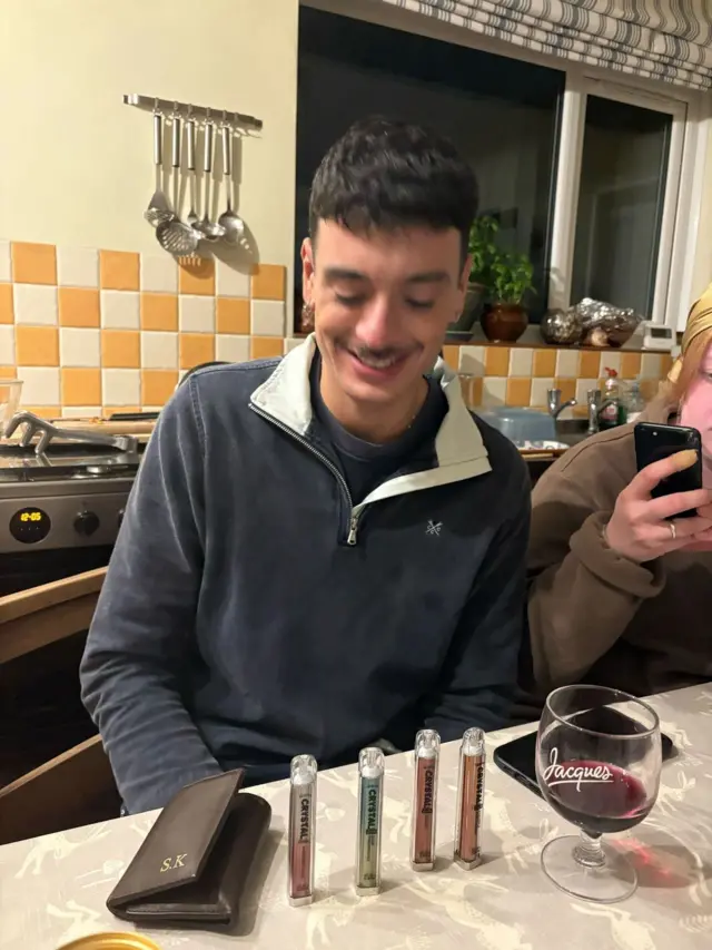 A man smiling sat at a kitchen table as he looks down at four vapes lined up on the table, alongside a glass of wine and a wallet