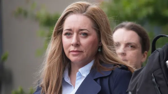 A red haired woman wearing a navy suit and blue shirt. A brown haired woman can be seen over her left shoulder.