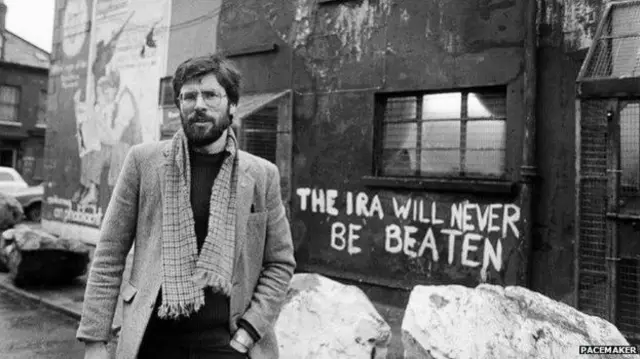 Gerry Adams standing in front of a wall with graffiti reading: "The IRA Will Never Be Beaten". He has think dark hair and beard, wearing glasses, a coat black jumper and scarf.