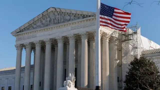 A fancy building exterior which has several large columns and a statue of someone sitting down, as well as a US flag on a pole in front of the building