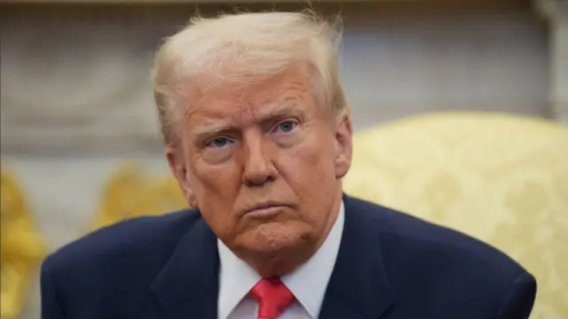 Close up of Donald Trump in a dark blue suit, white shirt and red tie as he sits on a yellow brocade chair inside the Oval Office