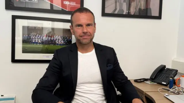 A man sits at a desk at smiles at the camera. He puts one hand on the desk which has a laptop on it. There's pictures on the wall behind him, on of a group picture