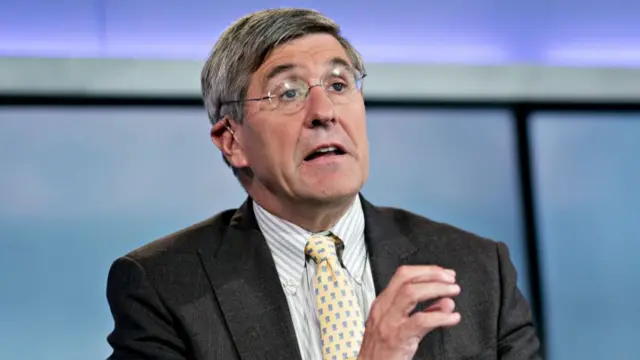 Stephen Moore (middle aged man with dark grey and white hair) mid-speech as he sits down in a black suit, striped shirt and pastel yellow and baby blue patterned tie