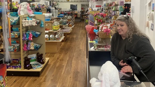 Mary Miller standing behind the counter of her shop. She's leaning on the counter wearing a black jumper, a pair of black reading glasses on top of her head, a bunny plush toy on the counter next to her