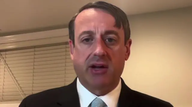 Jeffrey Schwab looks at the camera. He is wearing a black suit and a tie. He has brown hair.