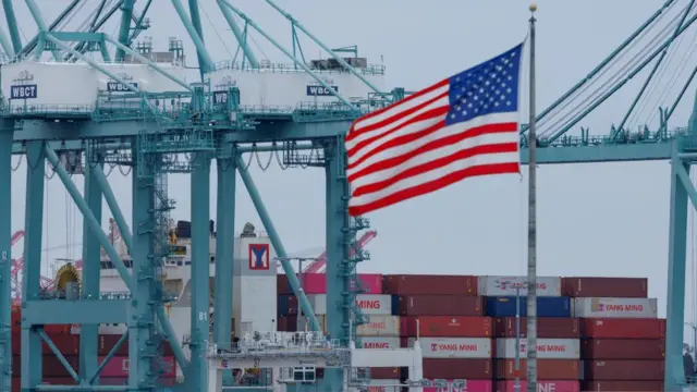 A stock photo of an American flag in front of a port, where shipping containers are piled high