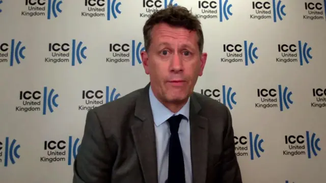 Chris Southworth (middle aged man with short brown hair) sits in a dark grey suit, light blue shirt and dark tie. Background is a wall covered in  the IIC United Kingdom logo