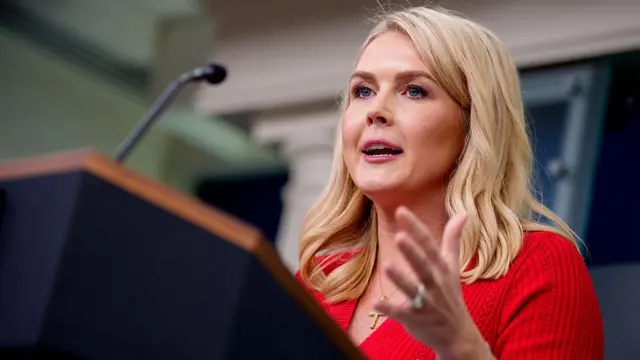 Karoline Leavitt speaks into a microphone. She is wearing a red dress. She has long blonde hair and blue eyes.