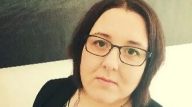 A selfie of a woman in front of a black and white background. She has short brown hair and wears glasses