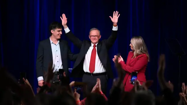 Prime Minister Anthony Albanese raises both arms and smiles on stage with his son Nathan and partner Jodie Haydon who smile toward Albanese.