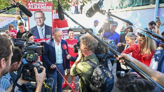 Anthony Albanese speakers to press outside his local polling station.  He is surrounded by photographers, journalists and camera operators.