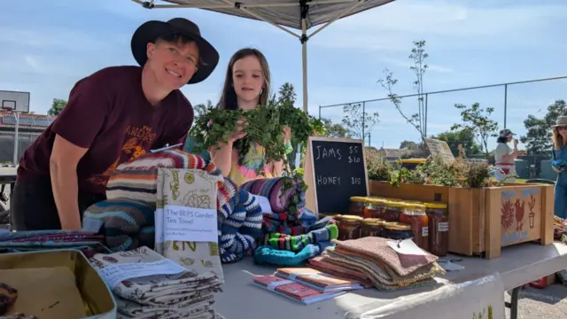 Annie Gehling in a large brown hat bending down and posing for the camera from behind her booth selling cloths and jars. Beside her is a young girl with long hair.