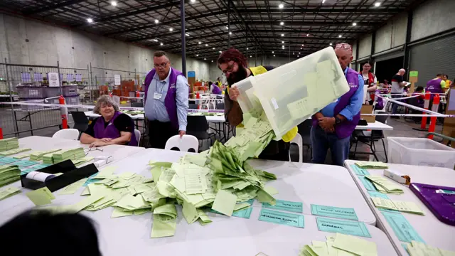 An Australian Electoral Commission staff member dumps a box of votes onto a table, as three of his coworkers look on from the side