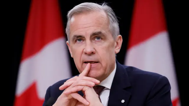 Mark Carney sits with two flags behind him, wearing a dark blue suit and red tie.