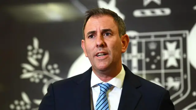 Australian Treasurer Jim Chalmers speaks to the media wearing a black suit, white shirt and blue striped tie.