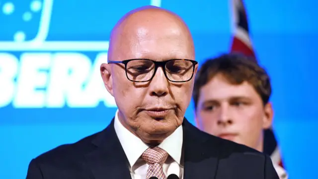 Peter Dutton lowers his gaze. He's wearing a suit. Behind him is his son.