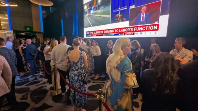 People dressed in formal attire stand in two lines in front of stanchions in a ballroom. Television news is projected on the wall behind them.