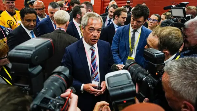 Nigel Farage in blue suit talking to media, surrounded by cameras with people wearing lanyards stood behind him.