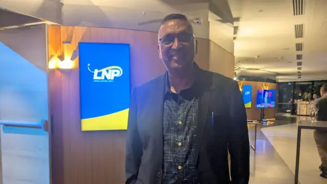 Jitendra Prasad has his hands in his pockets as he smiles for the camera. He is wearing a black suit and standing in front of a screen with the LNP logo