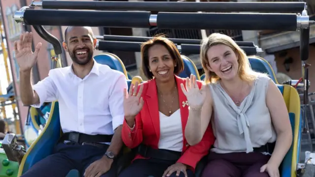 Munira Wilson sat on a rollercoaster with two people either side of her, she's wearing a red blazer and waving at the camera.