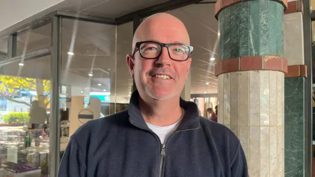 A man stands in front of a polling station in a dark blue zip up and glasses.
