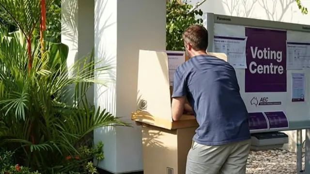 A man votes in person in Bali.  In the foreground you can see tropical plants next to the ballot booth.