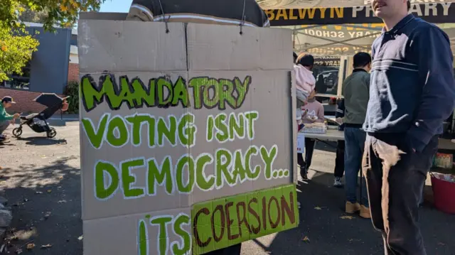 A man wears a sandwich board protesting against Australia’s compulsory voting. The message reads Mandatory voting isn't democracy… it's coercion” on the back.