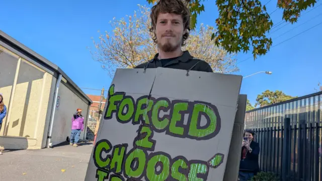 A man wears a sandwich board protesting against Australia’s compulsory voting, “Forced 2 choose, I Refuse” on the front