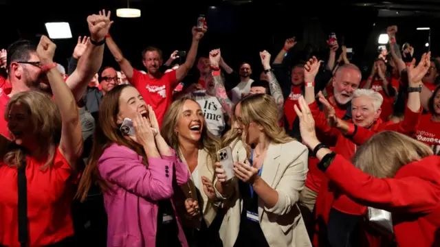 Supporters of Australia's Prime Minister Anthony Albanese react at a Labor party election night event. Many smile and raise their hands, with most dressed in red.