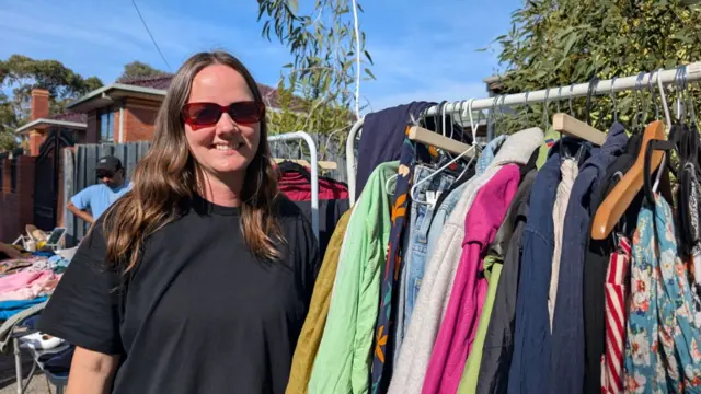 Tash Clough smiling at the camera, wearing a black t-shirt and red sunglasses. She's standing beside a rack of colourful clothes.