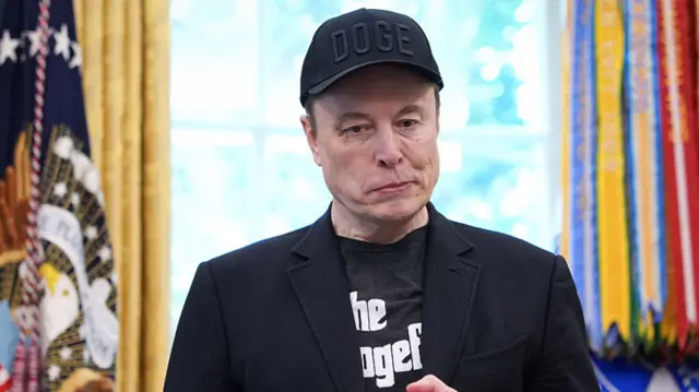 Musk wears a hat reading Doge and a black top reading The Dogefather in the Oval Office.