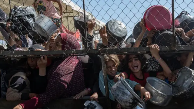 A crowd of people holding empty metal pots stand on the other side of a fence, some with anguished faces as the crowd appears to push forward