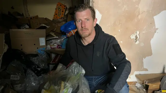 A man squatting on the floor looking into the camera surrounded by rubbish in a run down room. He look serious