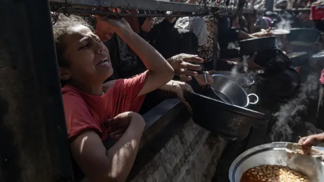 A child among the crowd waiting for food, with a look of distress on their face. Empty pots are held out behind them, as someone on the other side of the fence holds a full pot of beans to be handed out