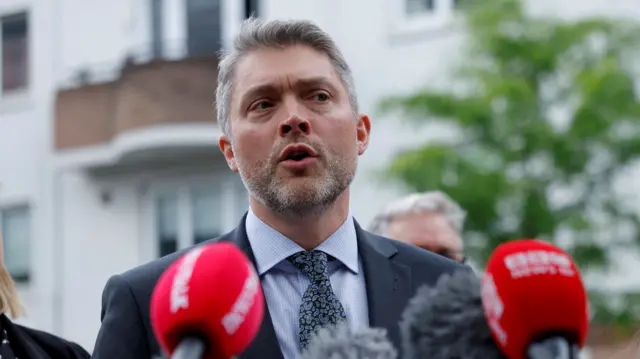 A man with short grey hair and beard is talking in front of two red microphones